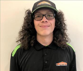 Male with long curly black hair wearing a SERVPRO hat and shirt.  Shirt has green on shoulders, he is wearing glasses.